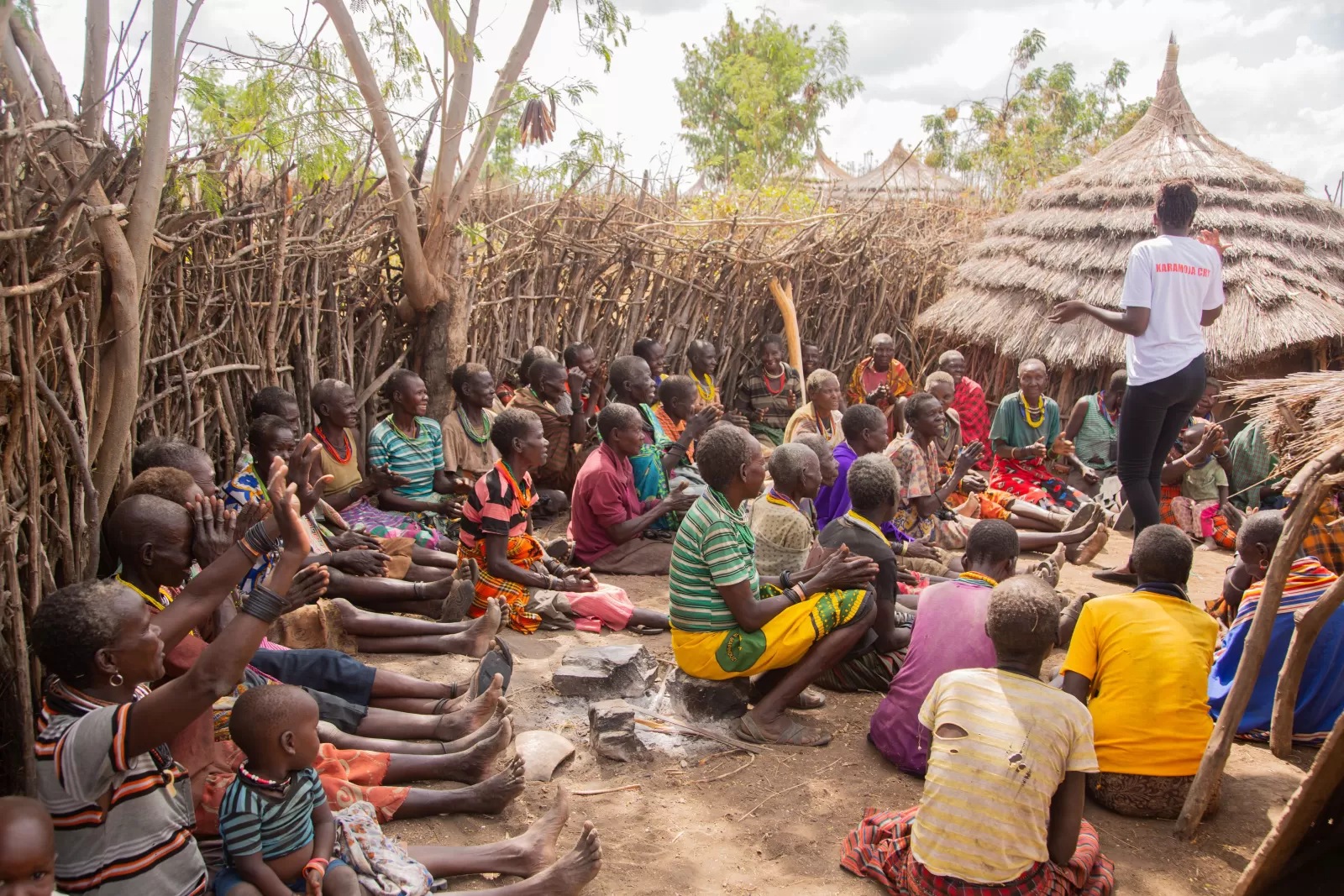A diverse group of adults and children gather under a tree for a community meeting outdoors.
